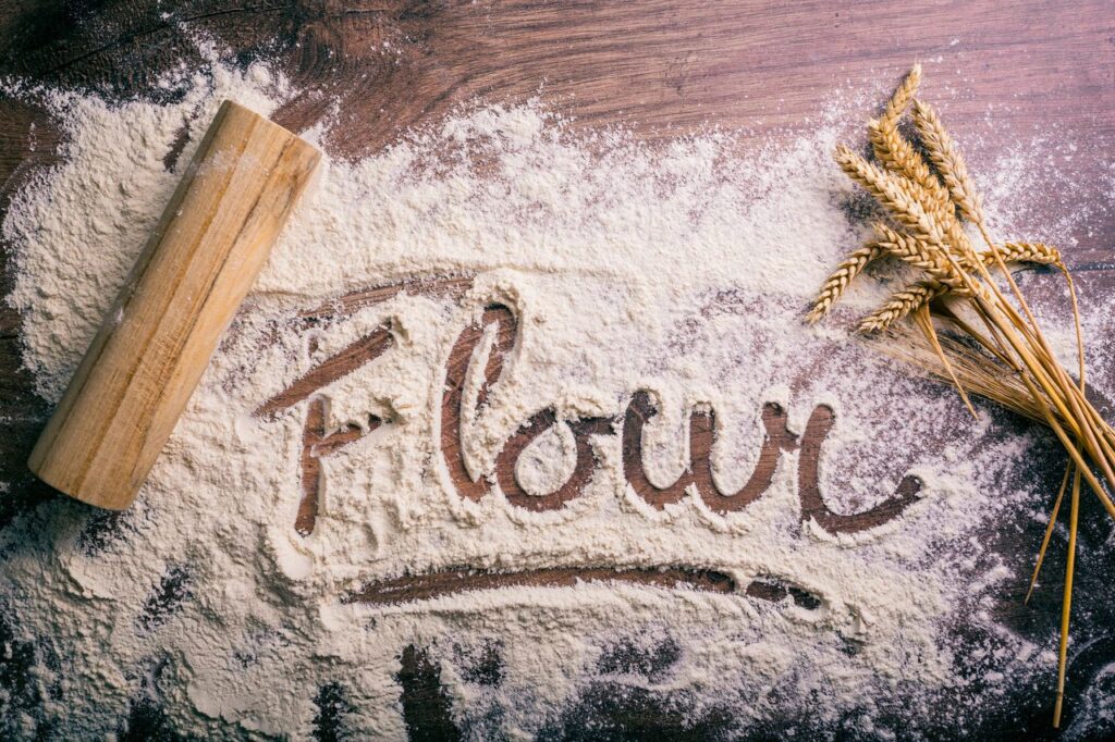 Flour scattered on a wooden table spelling 'flour,' with a rolling pin and wheat stalks nearby.